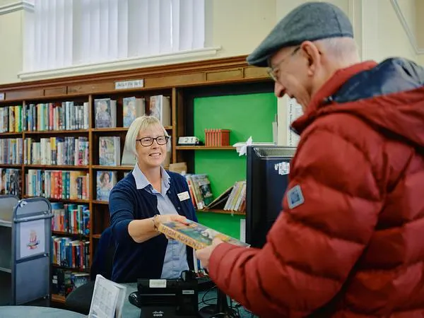 Library staff member handing a book to an older man in a red jacket at a library counter.