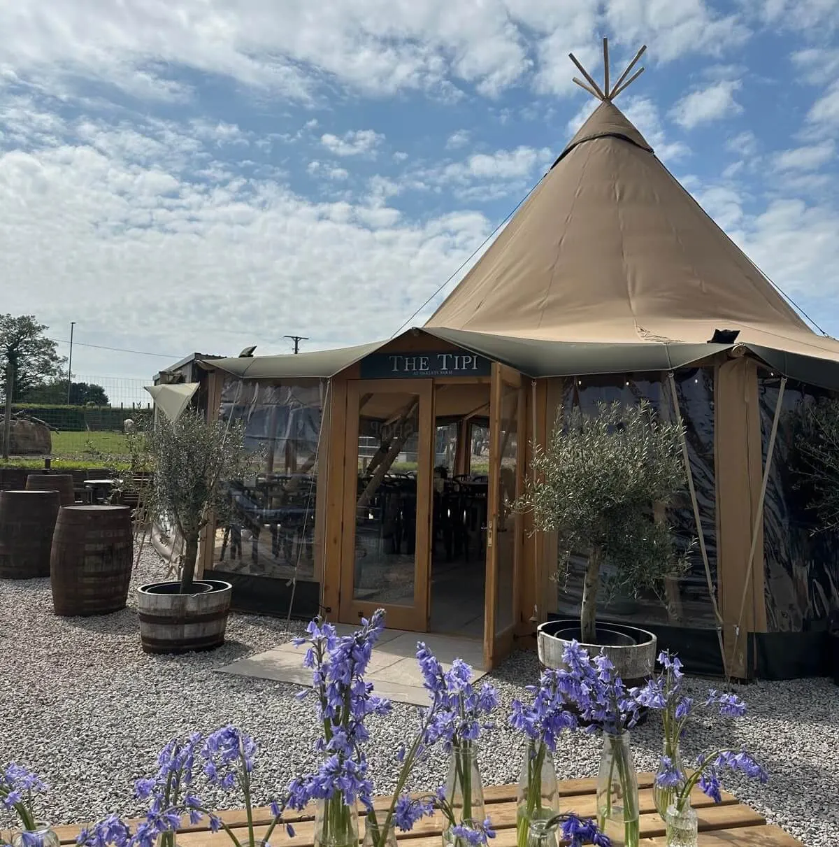 Glamping venue with a large beige tipi-style tent structure featuring wooden entrance doors, surrounded by purple flowers in vases and rustic barrels on gravel ground.