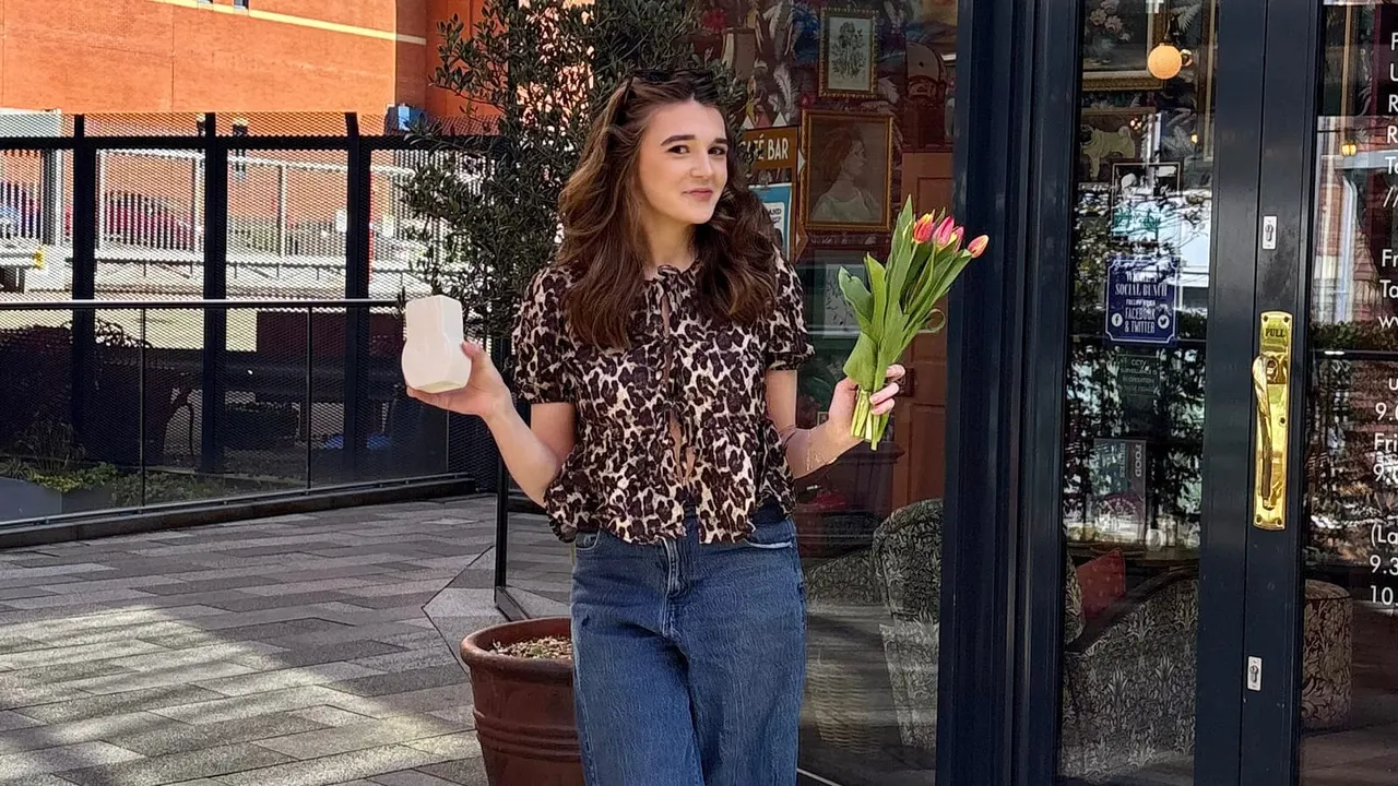Woman holding a drink and tulips standing outside a café storefront on a paved courtyard.