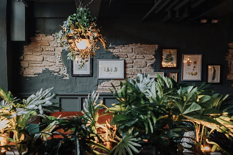 Interior décor featuring exposed brick wall with framed botanical artwork, hanging golden wreath with greenery, and potted plants in foreground.