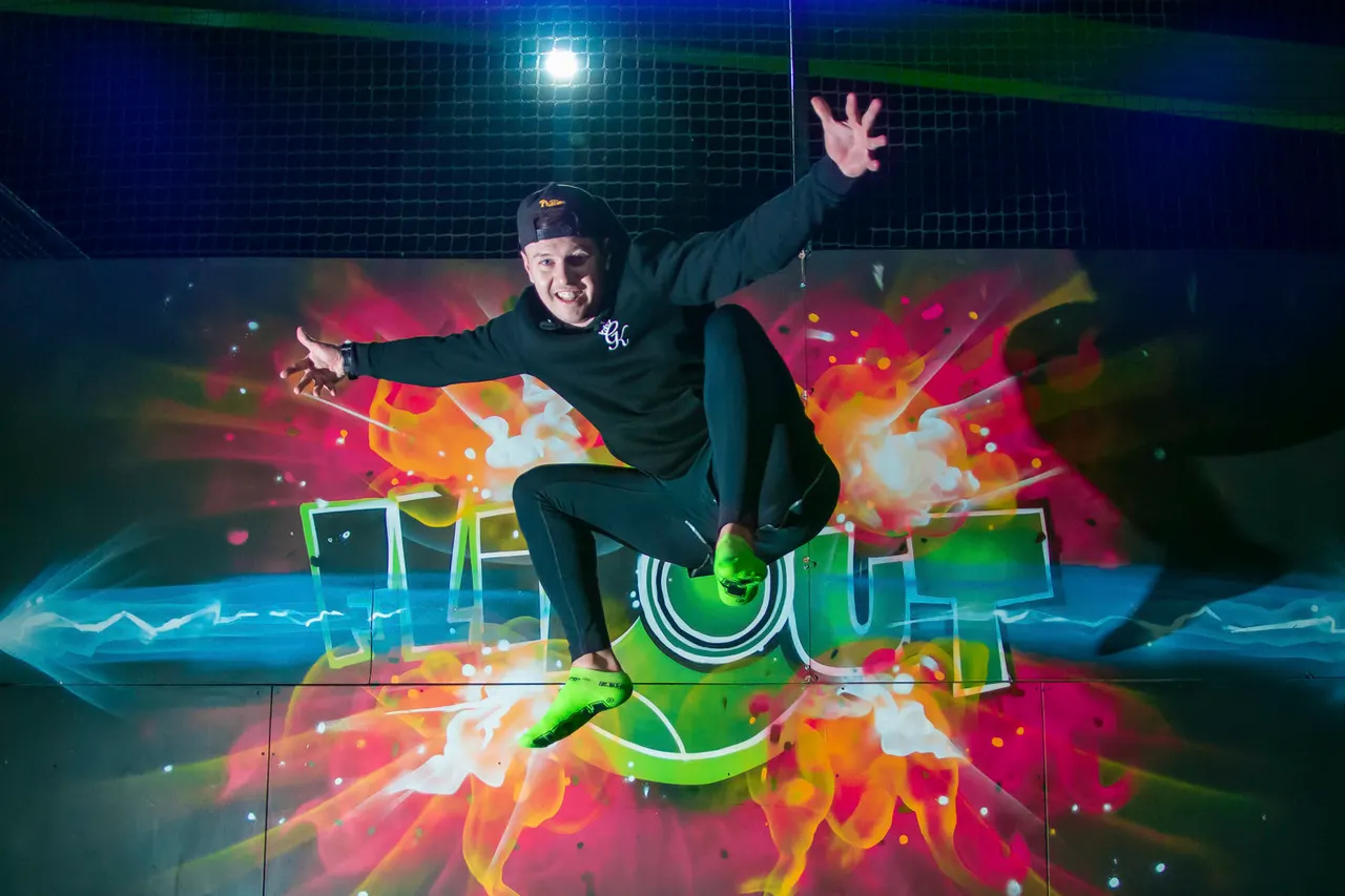 Person indoor skydiving with arms outstretched, smiling at camera against colourful projection backdrop.