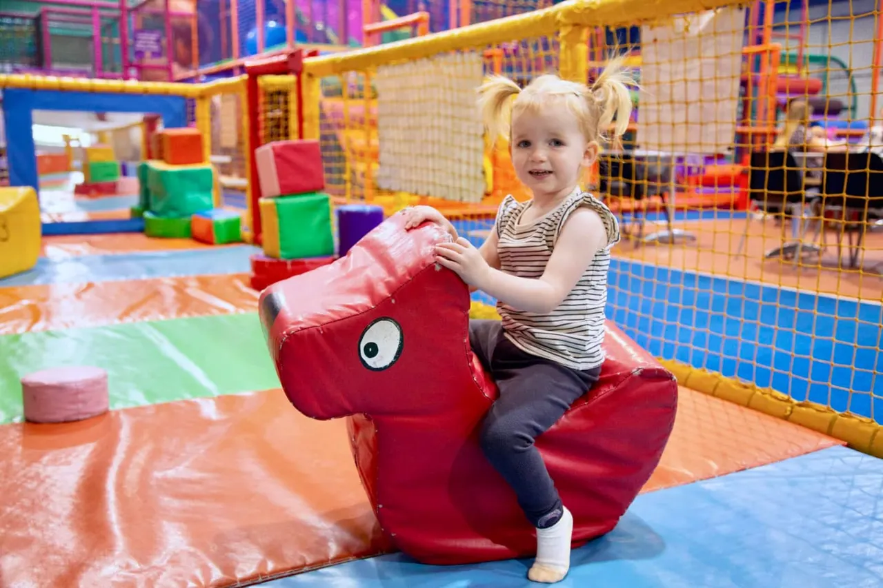 A young child with blonde pigtails rides a red inflatable toy in a colourful indoor soft play area.