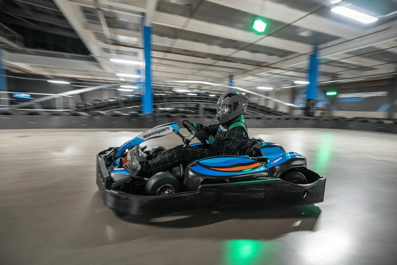 A person in a helmet and green jacket races a blue and black go-kart inside an indoor arena with blue support pillars and green exit lights.