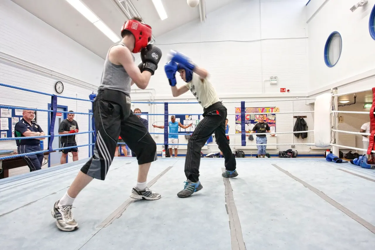 Two boxers in a ring wearing protective headgear and gloves during a sparring match, with spectators watching.