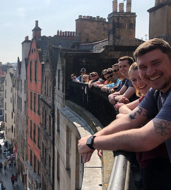 Visitors lean on a railing on a castle rooftop terrace, overlooking historic stone buildings and a street below.