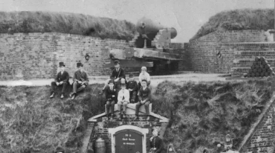 Group of visitors posed on cannon at brick fortification with steam locomotive in background.