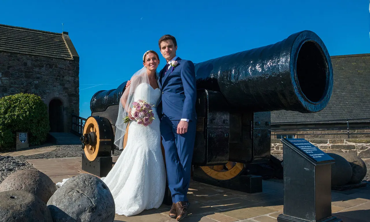 Bride and groom posing together beside a large historic cannon at a fortified gatehouse under clear blue skies.