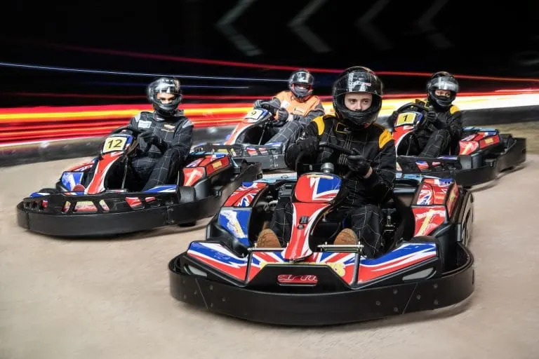 Multiple people in racing suits and helmets driving red and blue go-karts on an indoor track with neon lighting.