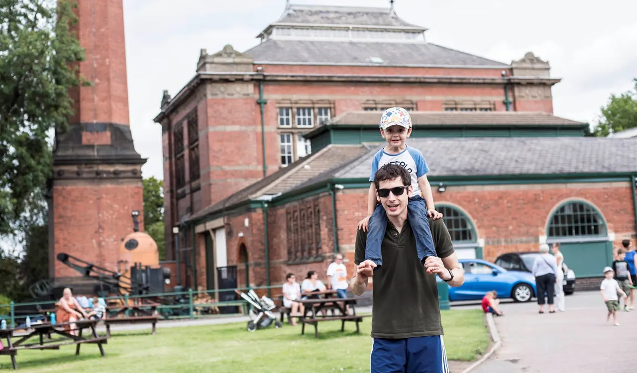 Man carrying young child on shoulders in front of red brick Victorian building with families relaxing on grass.