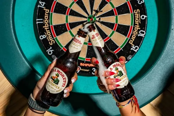 Two people holding beer bottles in front of a dartboard with darts.