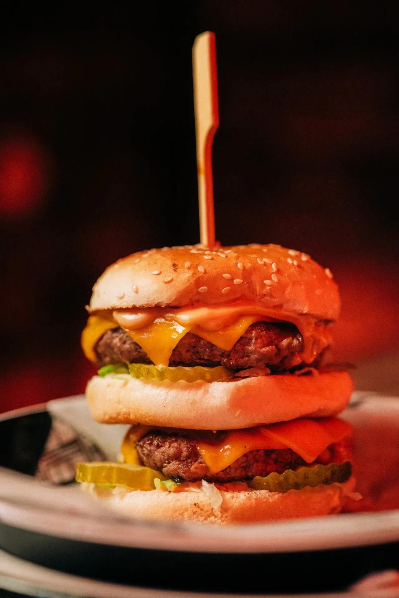 A stacked double cheeseburger with sesame seed buns, melted cheese and pickle slices, held together with a wooden skewer on a plate.