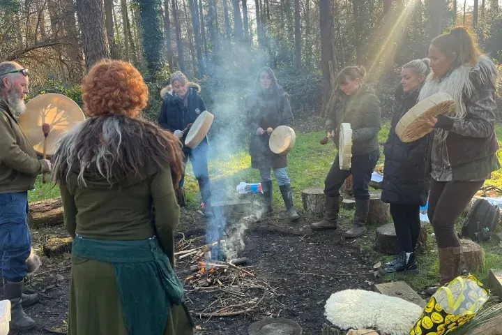 Group of people gathered in a woodland clearing around a central fire, holding drums and participating in a drumming circle activity.