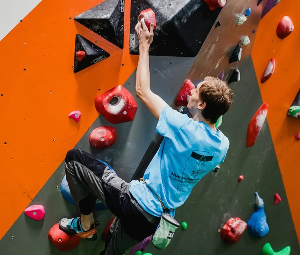 Person rock climbing on an indoor wall with coloured holds, viewed from above.