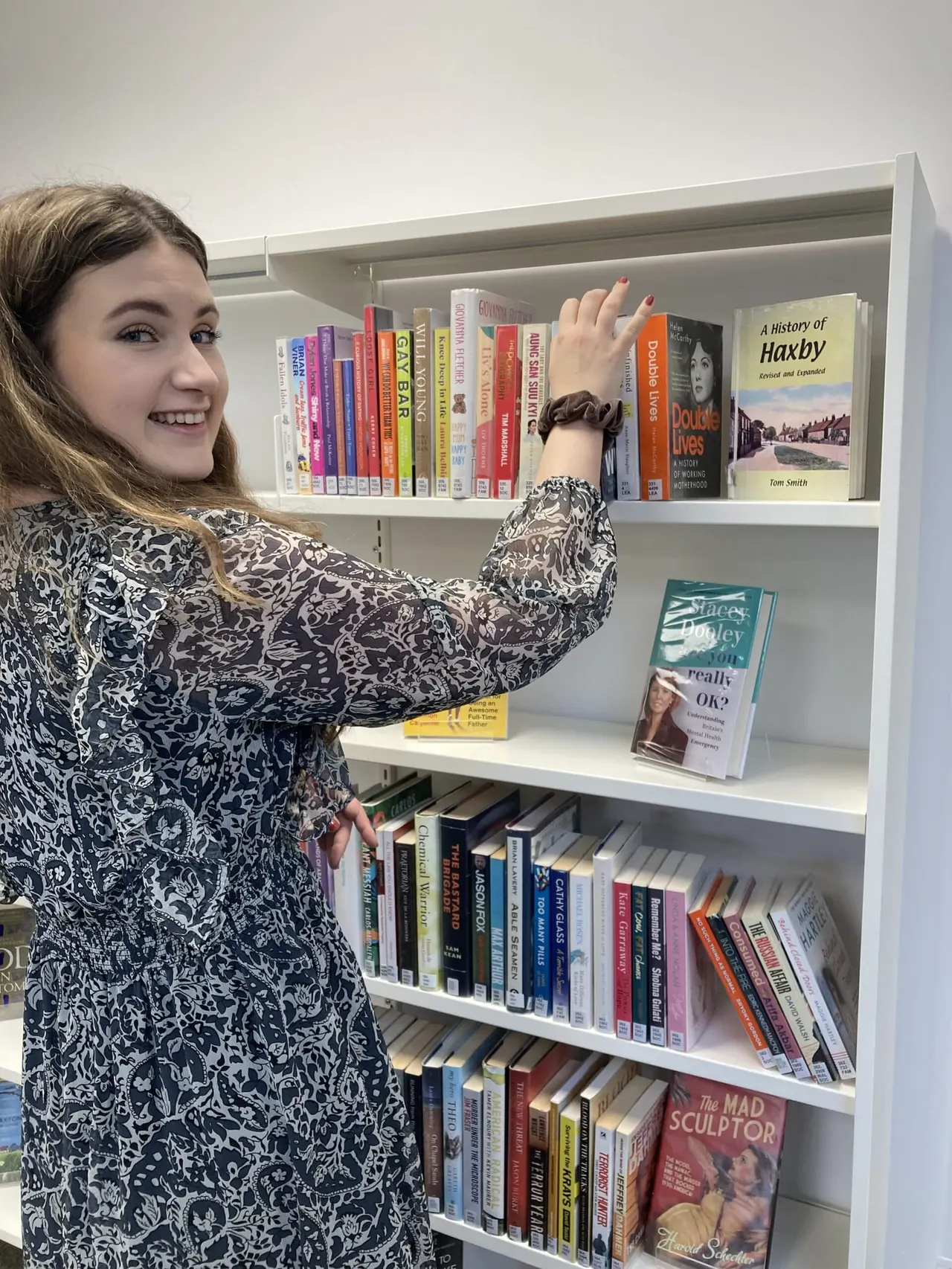 A young woman smiling whilst reaching for a book on a white shelving unit filled with colourful books.