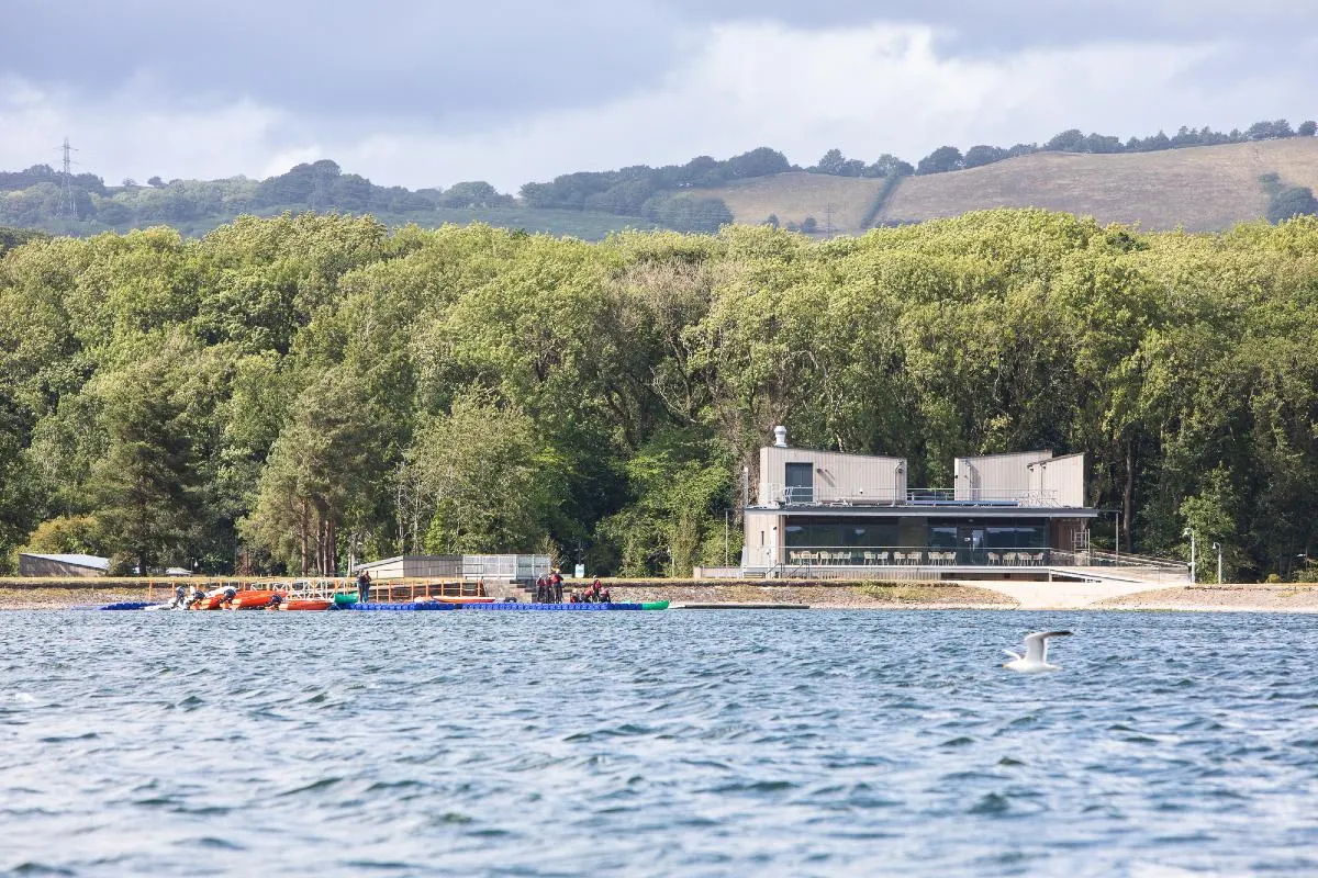 Waterside leisure facility with white modern building and colourful inflatables by the shore, forested hills beyond.