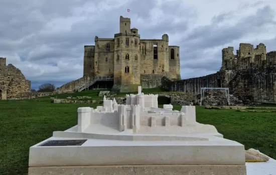 A white architectural model of Warkworth Castle sits on display on the castle grounds, with the actual stone castle keep and ruins visible behind it.