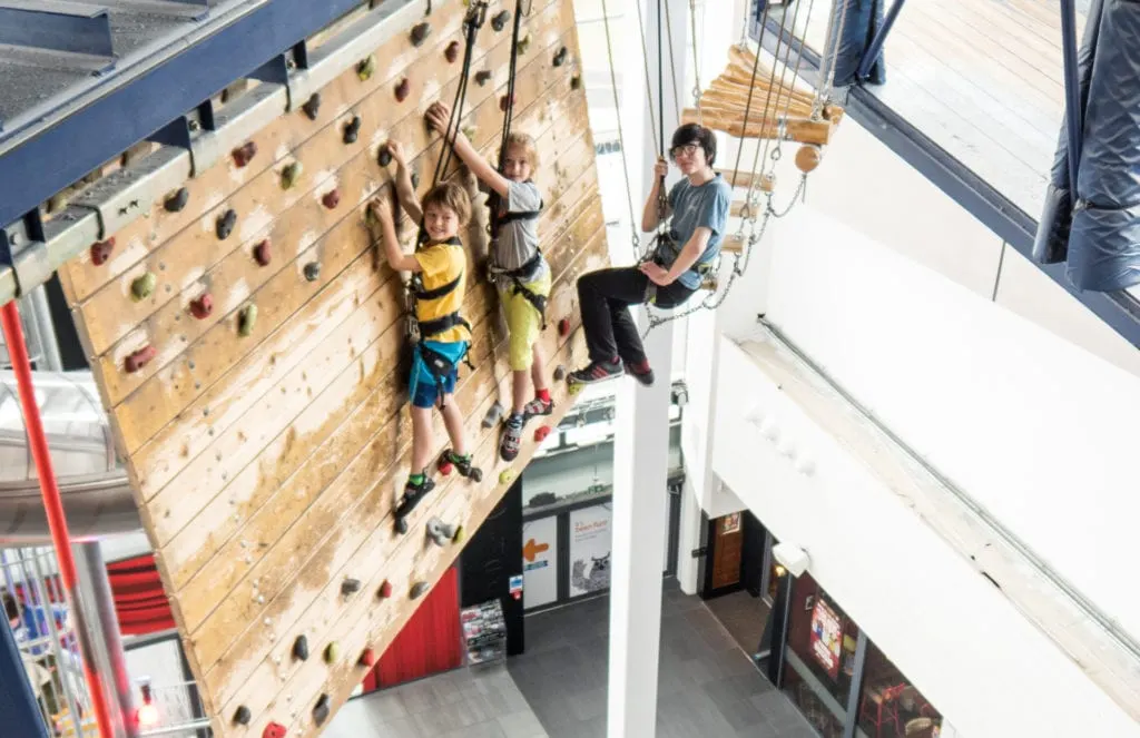Children and teenagers climbing an indoor rock wall with safety harnesses indoors.