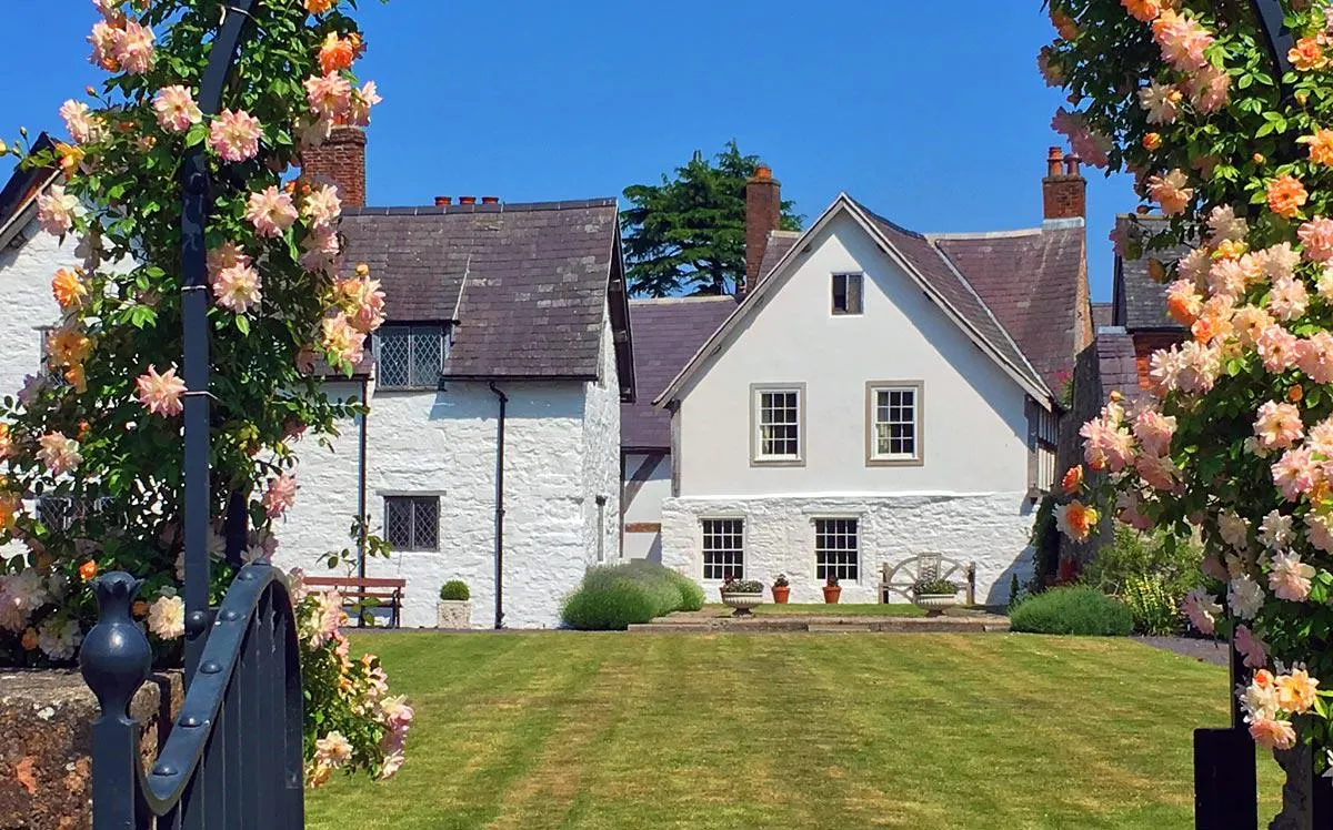 PHOTOGRAPH

Historic white-walled house with slate roof surrounded by manicured lawns and framed by climbing roses in bloom on either side.