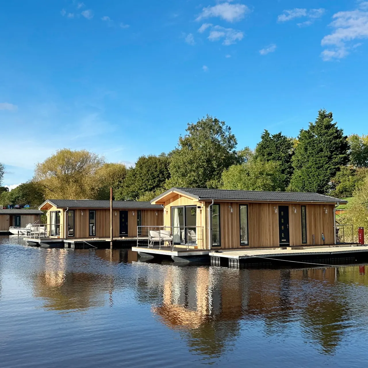 Modern wooden waterside lodges with decking moored on a river, surrounded by trees under blue sky.