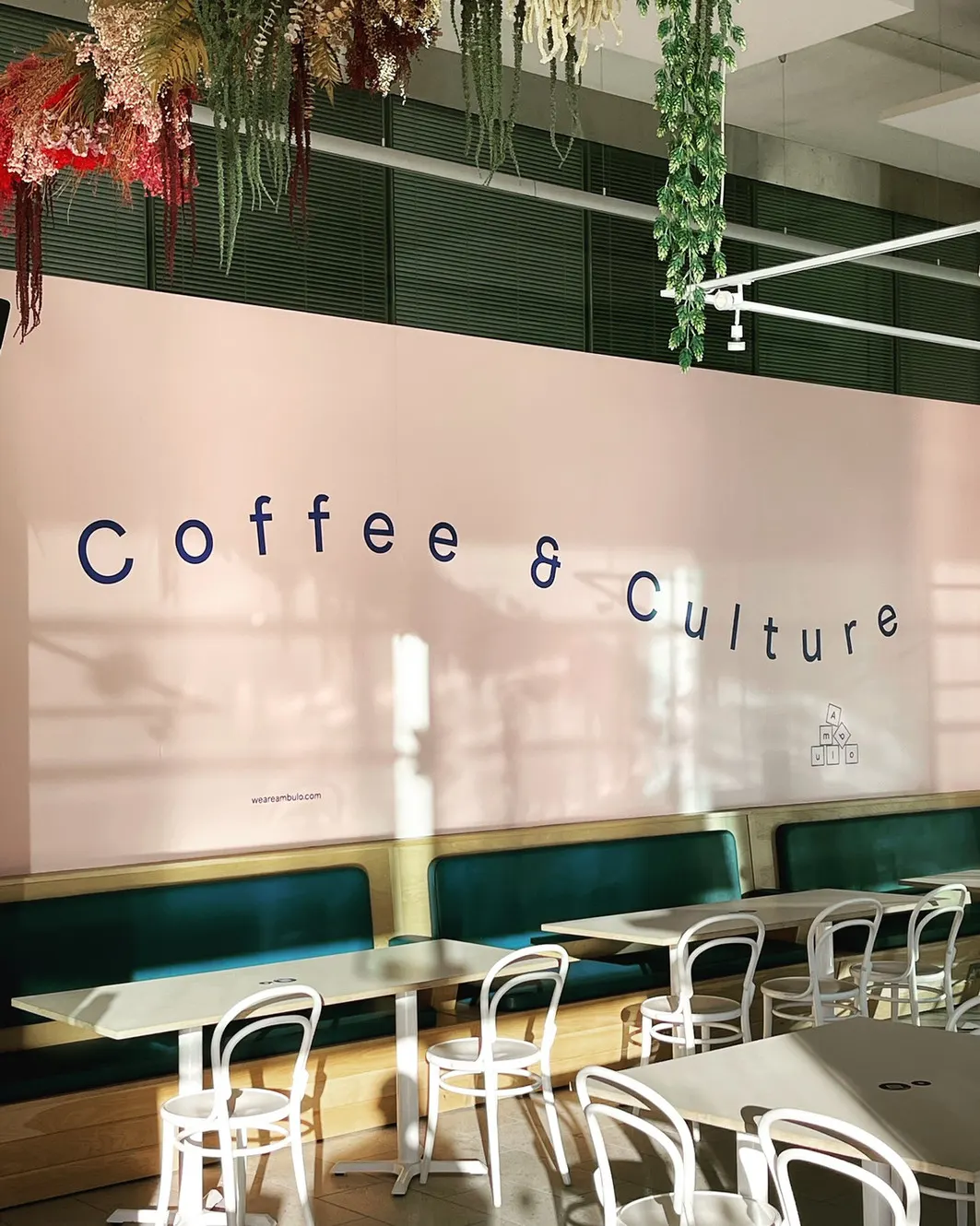 Modern café interior with white bentwood chairs, green-upholstered banquettes, and "coffee & culture" wall text with hanging plants.