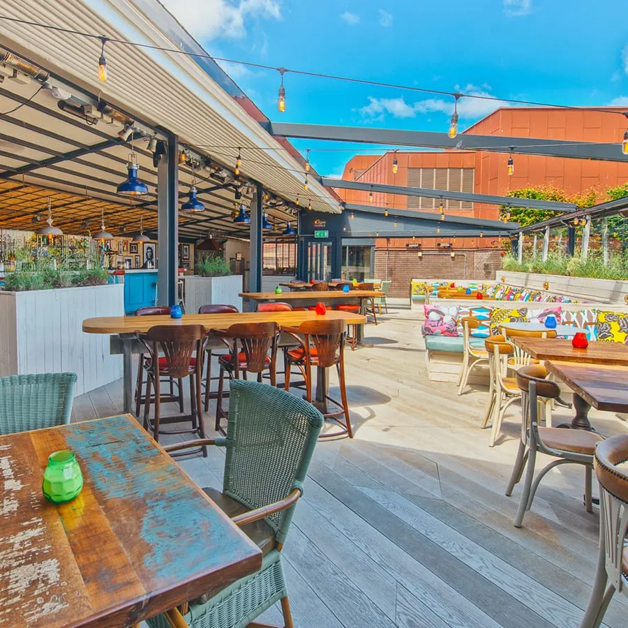 Spacious outdoor terrace with wooden tables, bar seating, and colourful décor under metal pergolas on a sunny day.