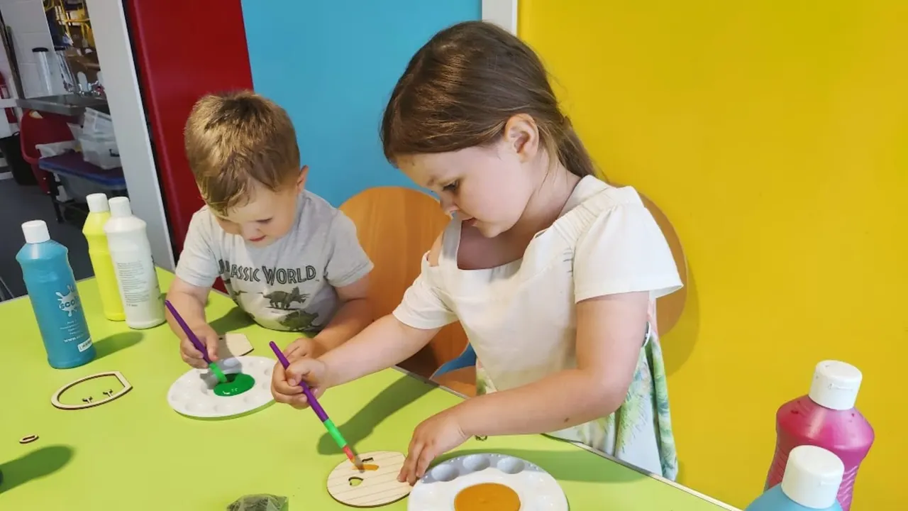 A young girl and toddler paint together at a bright yellow table with colourful paint bottles nearby.