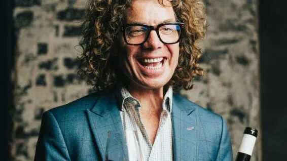 Man with curly hair and glasses wearing a blue blazer smiles at camera against a textured stone wall.