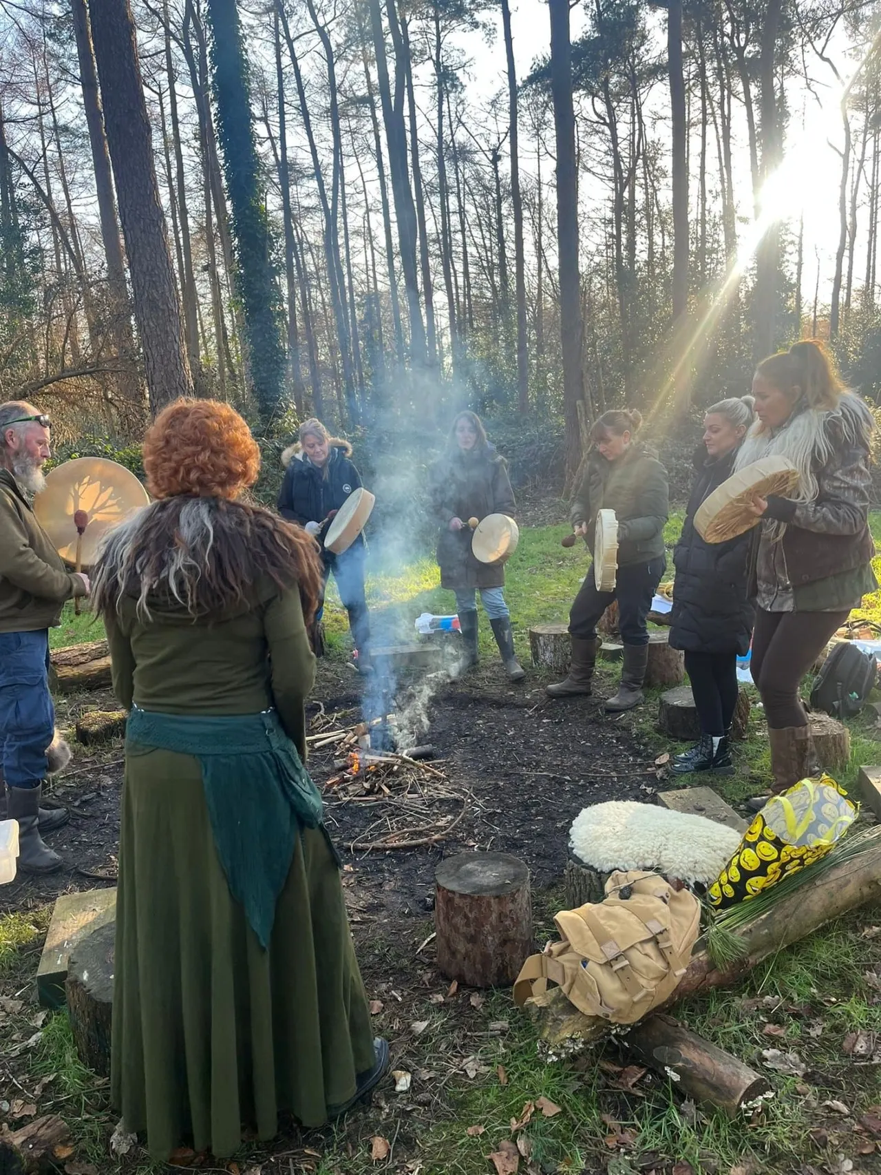Group gathering around a campfire in a woodland clearing, with participants holding drums and engaging in a music or drumming circle activity.