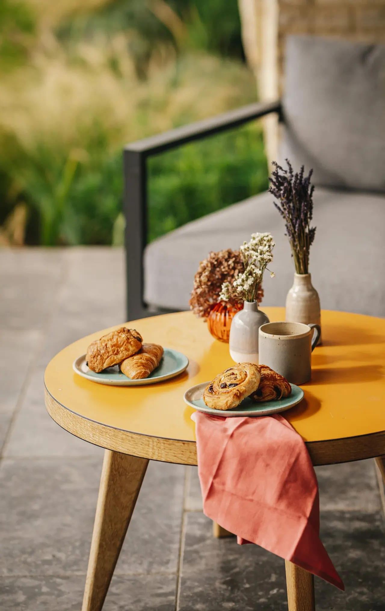 A yellow round table set with pastries, coffee, dried flowers in vases, and a pink napkin in an outdoor garden seating area.