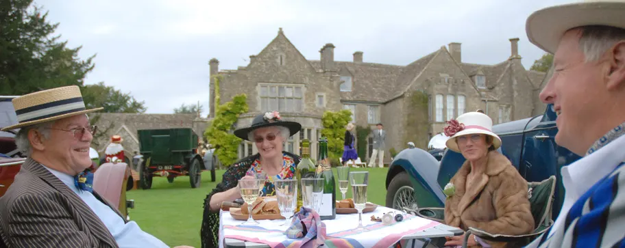 Guests in period costume enjoy a picnic lunch on the lawn in front of a stone manor house with vintage cars on display.