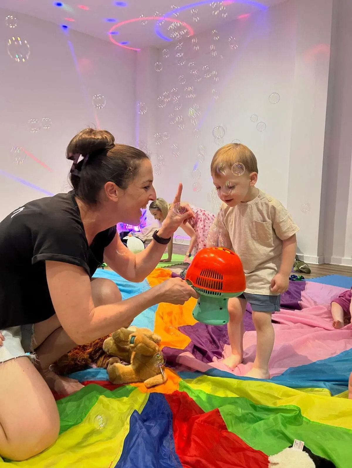 Adult and toddler playing together on colourful mats with bubbles and coloured lights in a sensory play room.