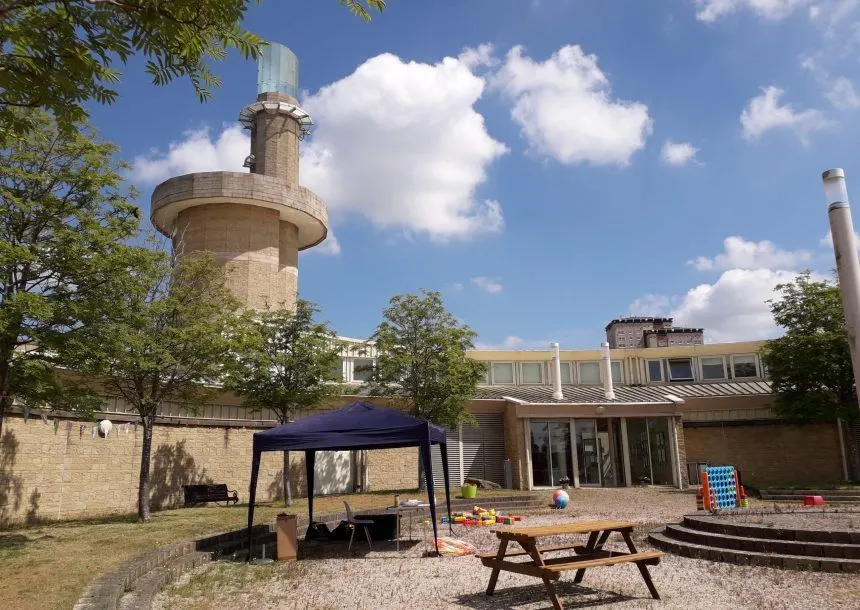 Outdoor play area with picnic tables and a blue canopy beside a brick building, with a historic stone tower and industrial chimney visible against a blue sky.