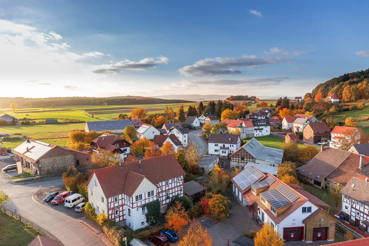 Aerial view of a rural village with traditional half-timbered houses, autumn foliage, green fields and farmland stretching to distant hills.