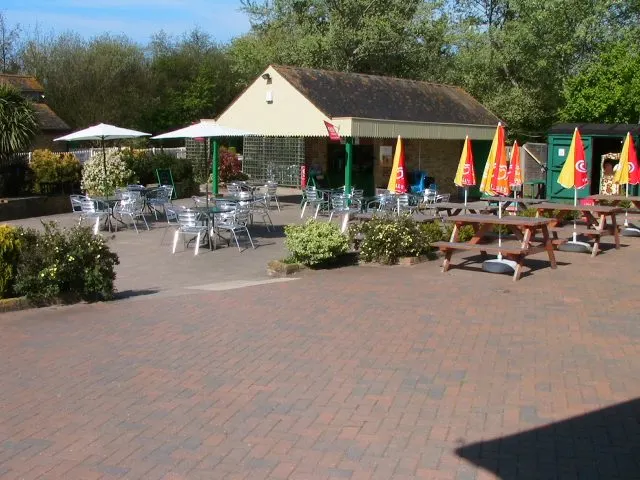 Outdoor leisure venue terrace with seating, colourful umbrellas, building and surrounding trees on a sunny day.