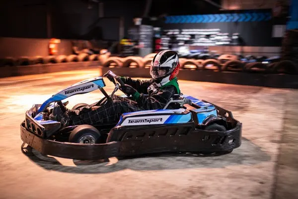 Person in racing helmet driving a blue and black go-kart on an indoor track with barriers.