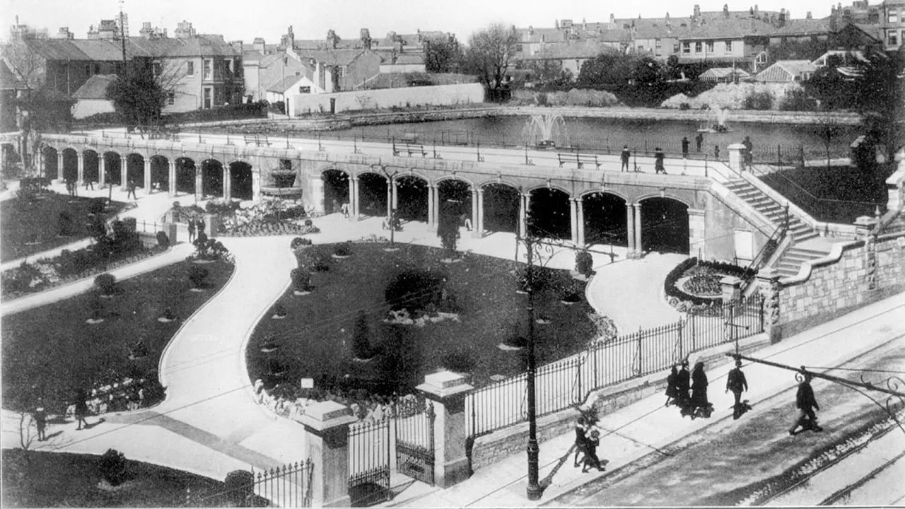 Historic open-air swimming pool complex with Art Deco changing pavilions, manicured gardens and visitors in 1930s seaside setting.