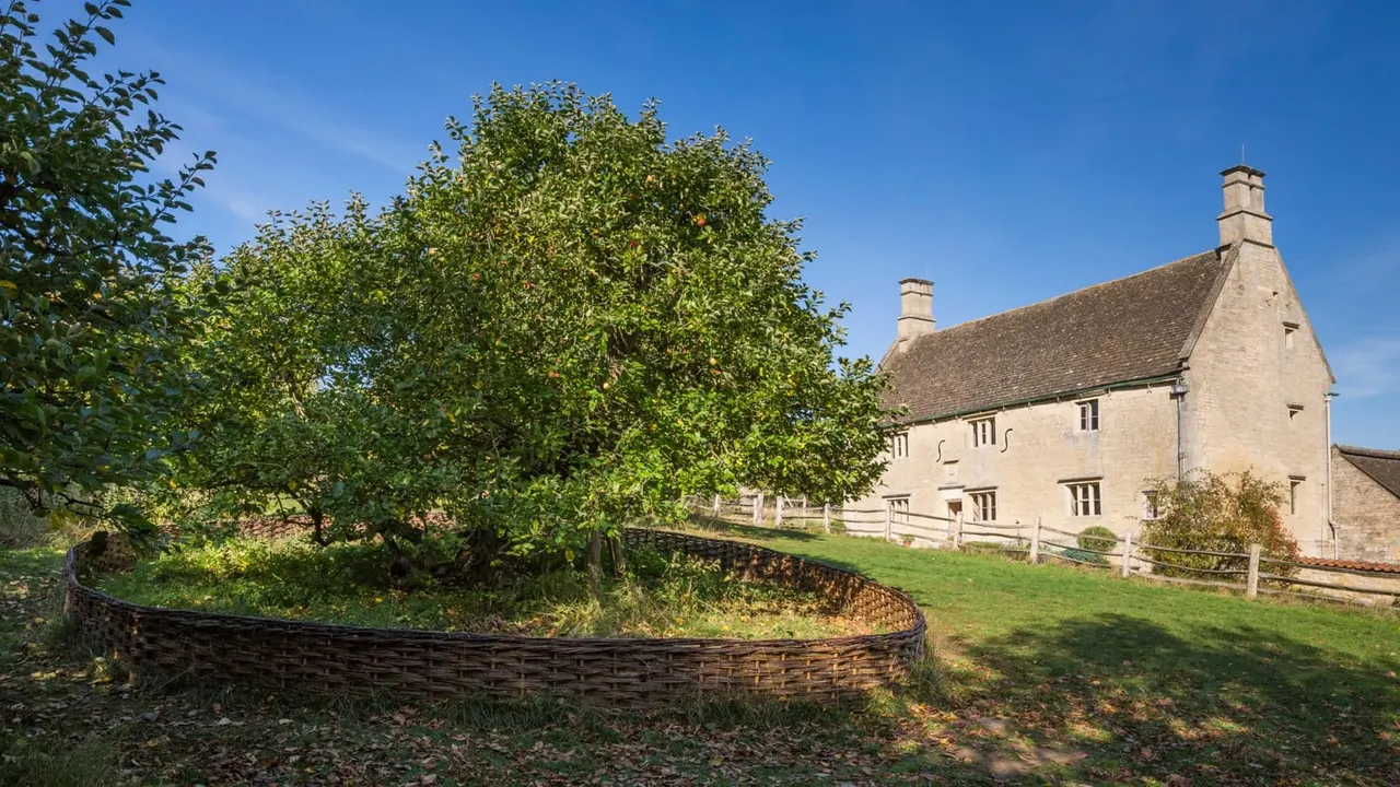 Stone farmhouse with wicker fencing and fruit trees in grassy grounds under clear blue sky.