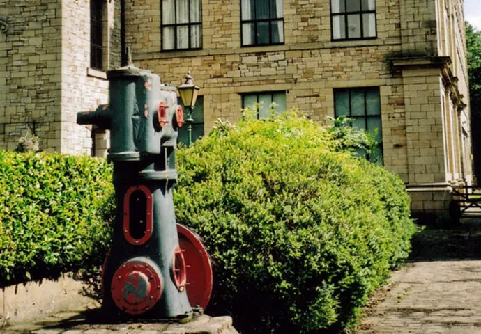 A red and green vintage steam engine boiler sits in a courtyard surrounded by clipped hedges against a stone building.
