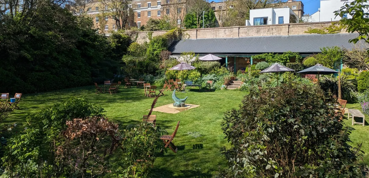 Spacious outdoor garden with lawn seating, parasols, and wooden chairs surrounded by hedges and period buildings.