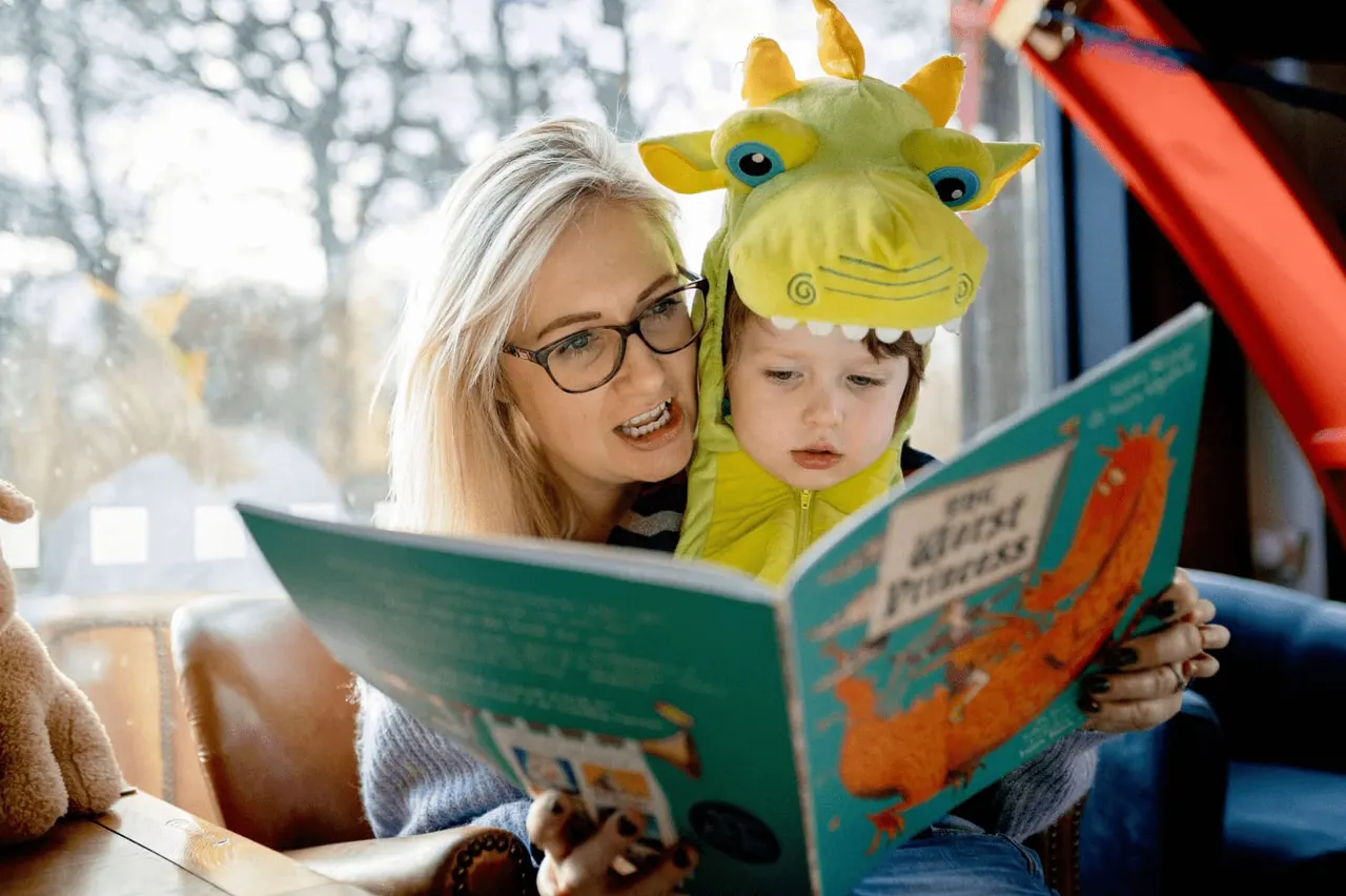 Adult and young child wearing a green monster hat reading a picture book together indoors.