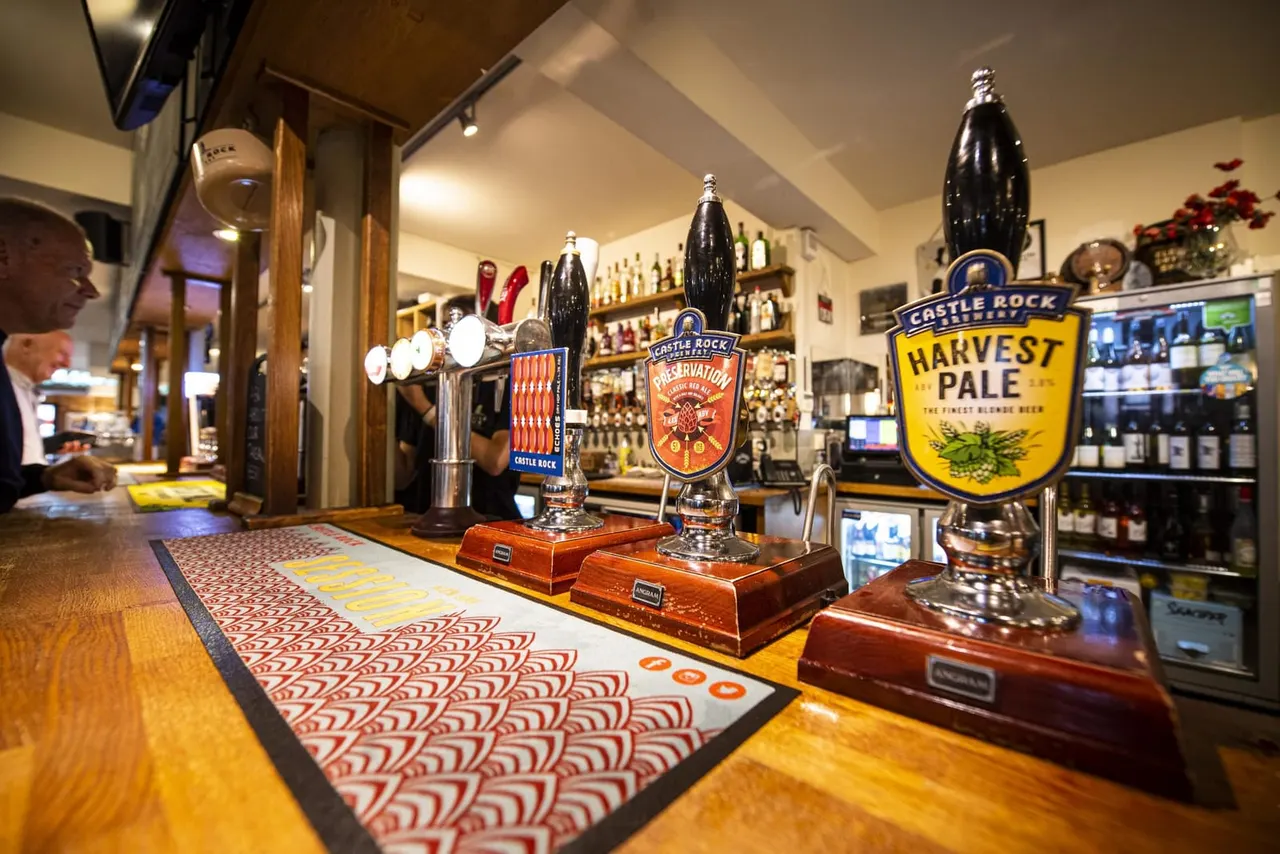 Beer pump handles and taps lined up on a pub bar counter, with bottle shelves and customers in the background.