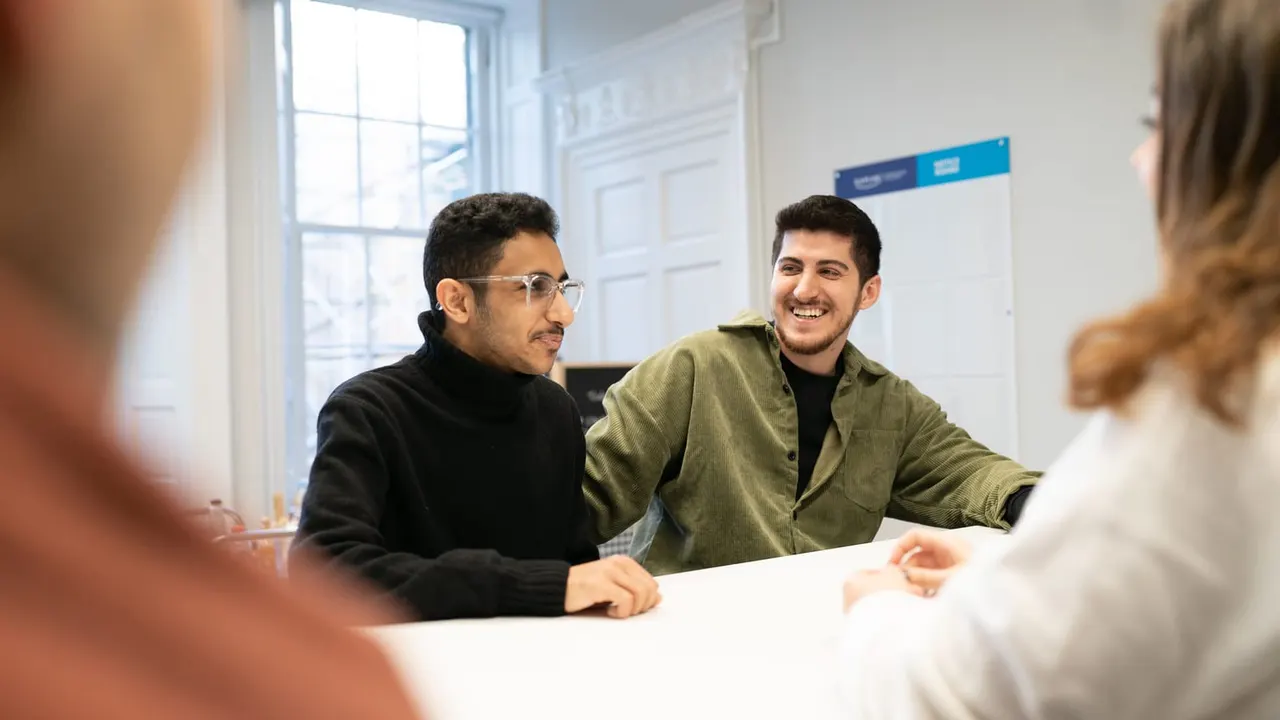 This is a PHOTOGRAPH.

Two young men sit at a table during a group discussion or lesson in a bright classroom with large windows.