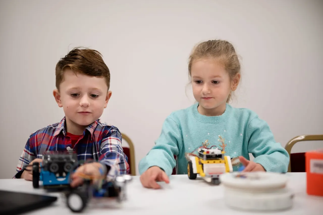 Two young children sitting at a table working with small robotic vehicles and electronic building components.
