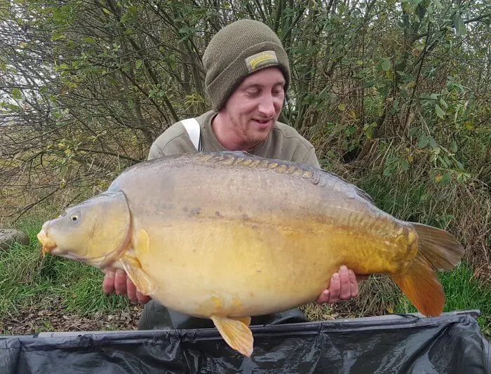 Man holding a large carp he has caught at a fishing venue, with trees in the background.