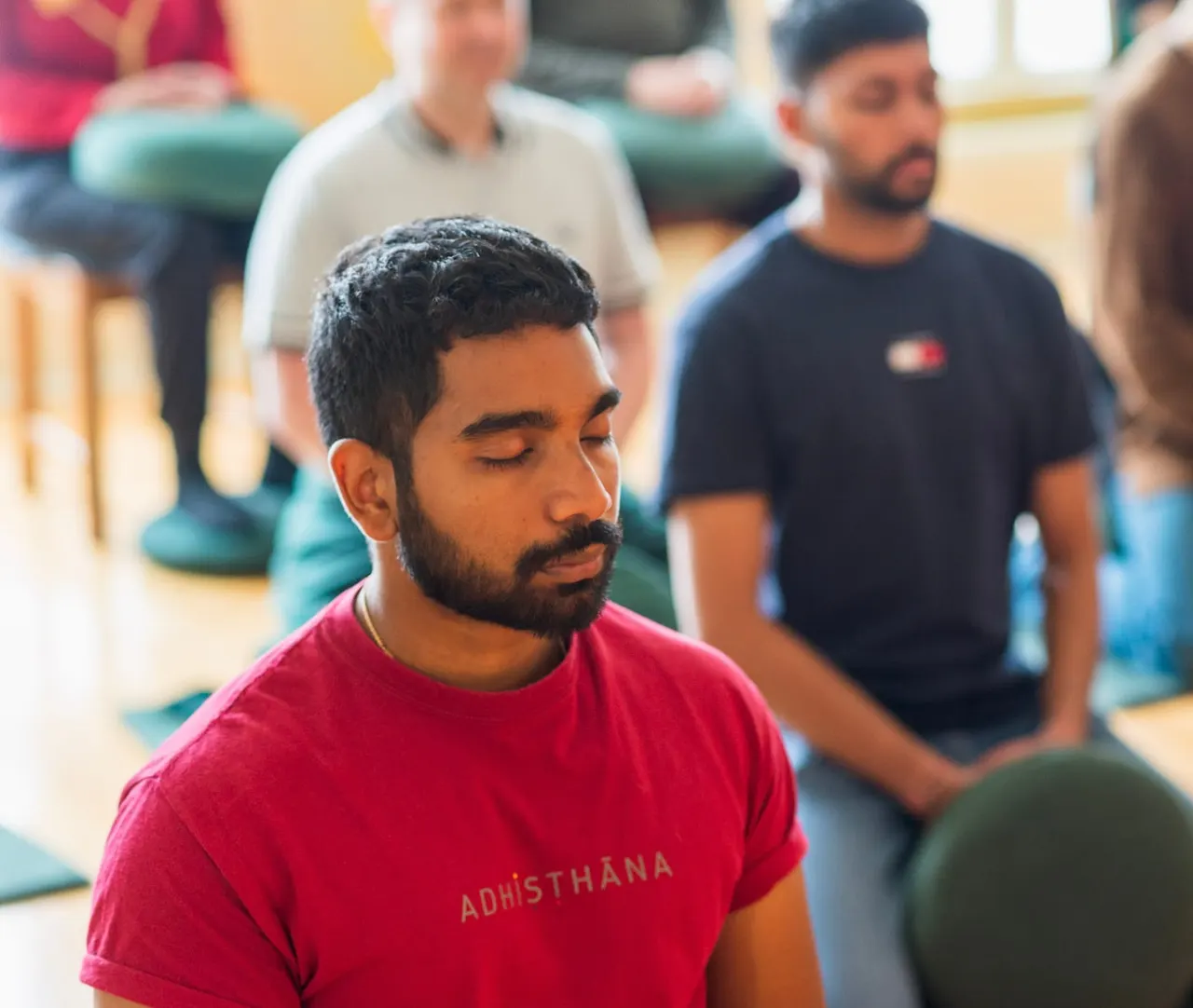 Man in red shirt with eyes closed during meditation session with other participants seated on mats indoors.