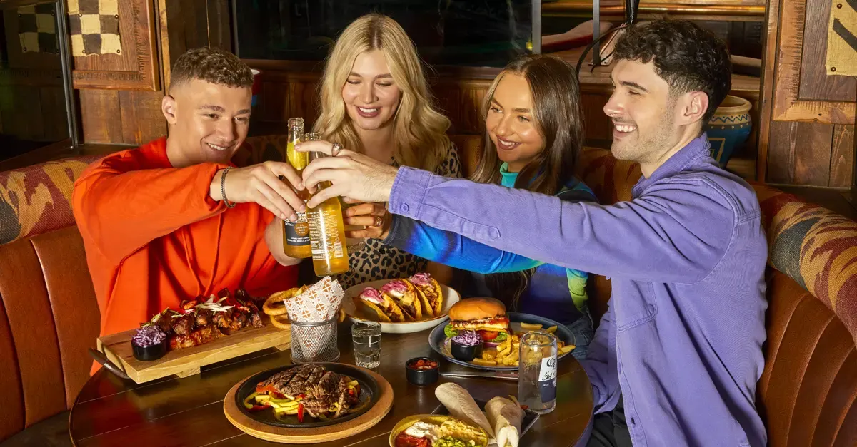 Four friends clinking glasses of beer during a meal with burgers, ribs and other food on the table.