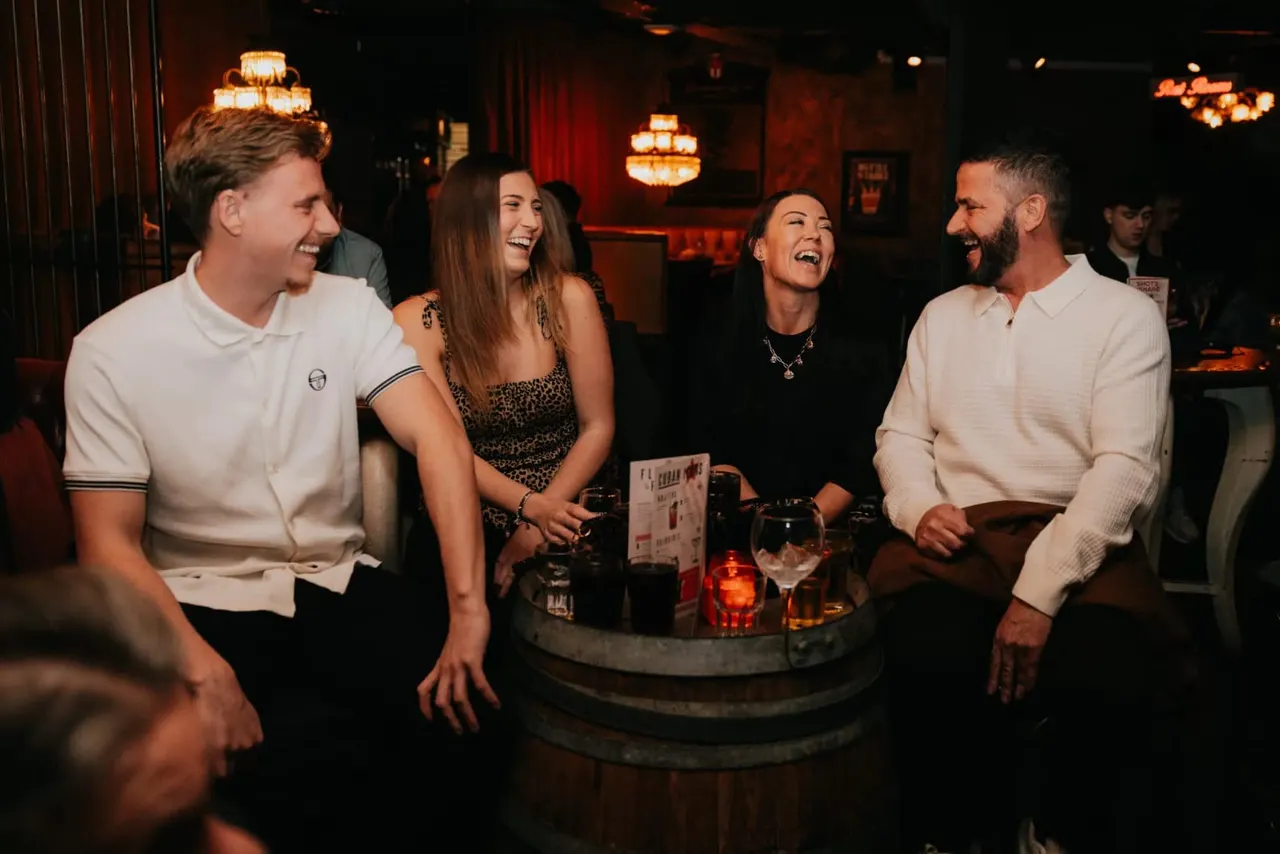 Four people laughing whilst standing at a barrel table with drinks in a dimly lit nightclub with warm orange lighting.