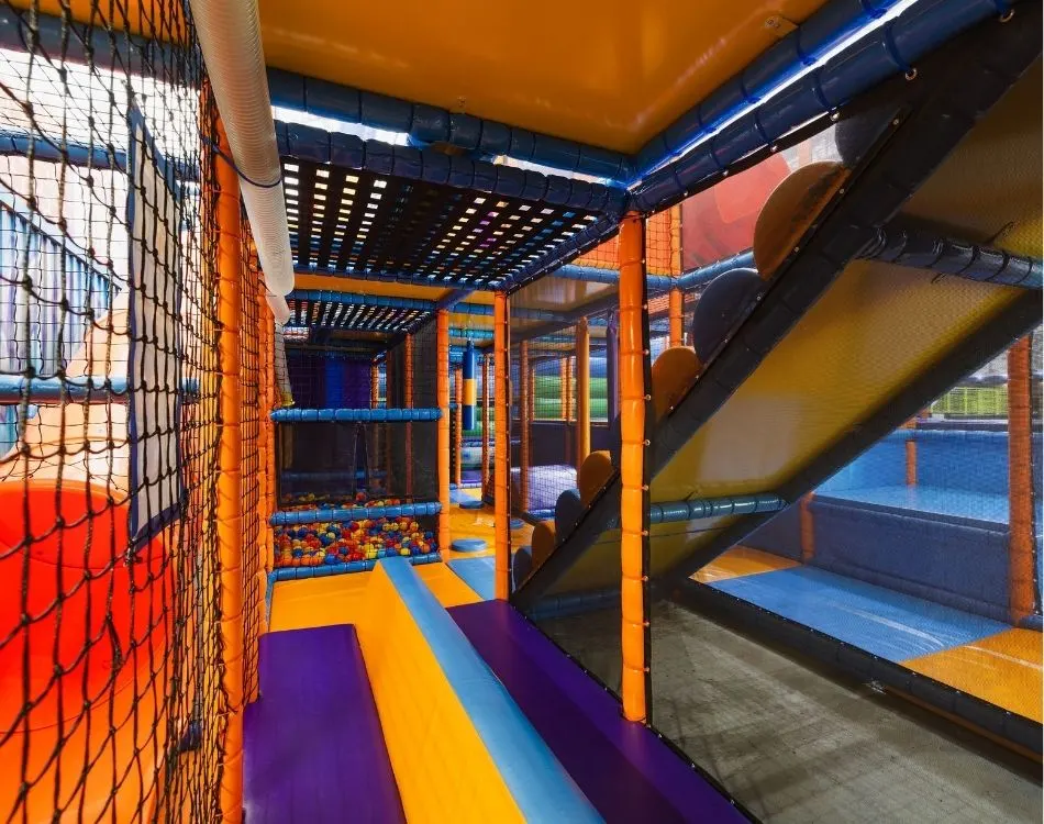 Interior of a colourful indoor play centre with safety netting, platforms, slides and ball pit areas.