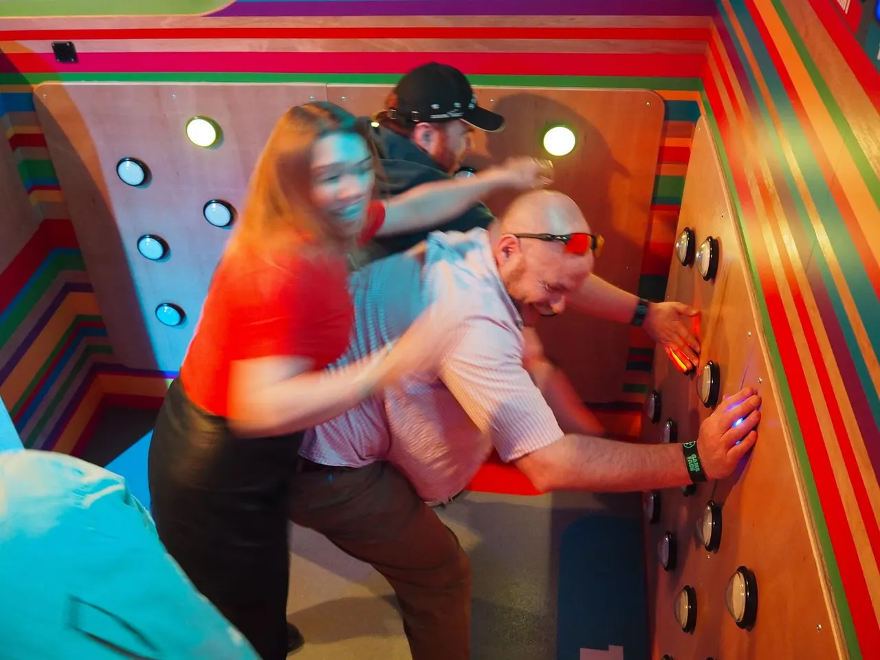 Three people playing an interactive arcade game in a brightly lit, colourfully striped game booth.