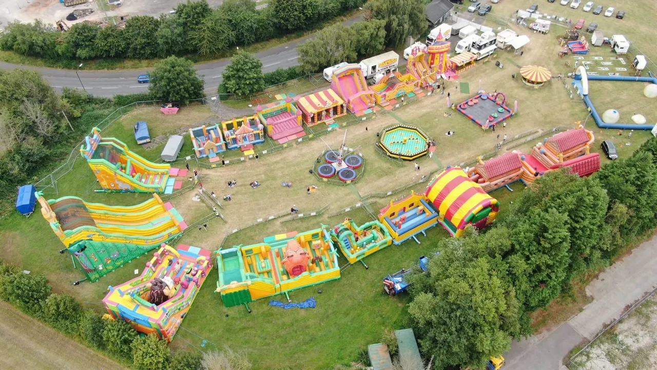 Aerial view of an inflatable fairground with colourful bouncy castles, slides and rides set up on a grass field surrounded by trees.